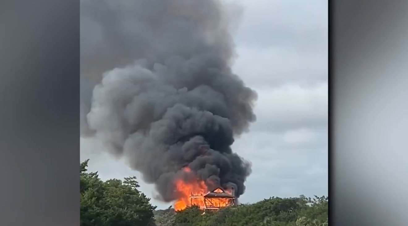 A house engulfed in flames emits thick black smoke against a cloudy sky, surrounded by greenery.