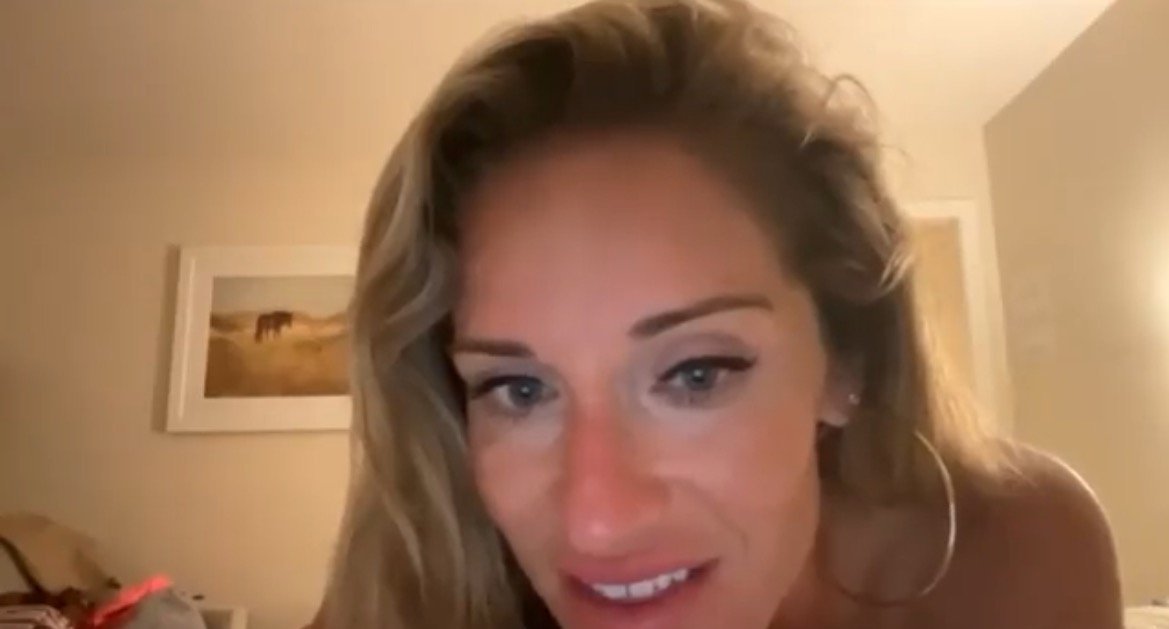 Close-up of a woman with long hair smiling while sitting in a softly lit room, with a framed artwork and personal items in the background.