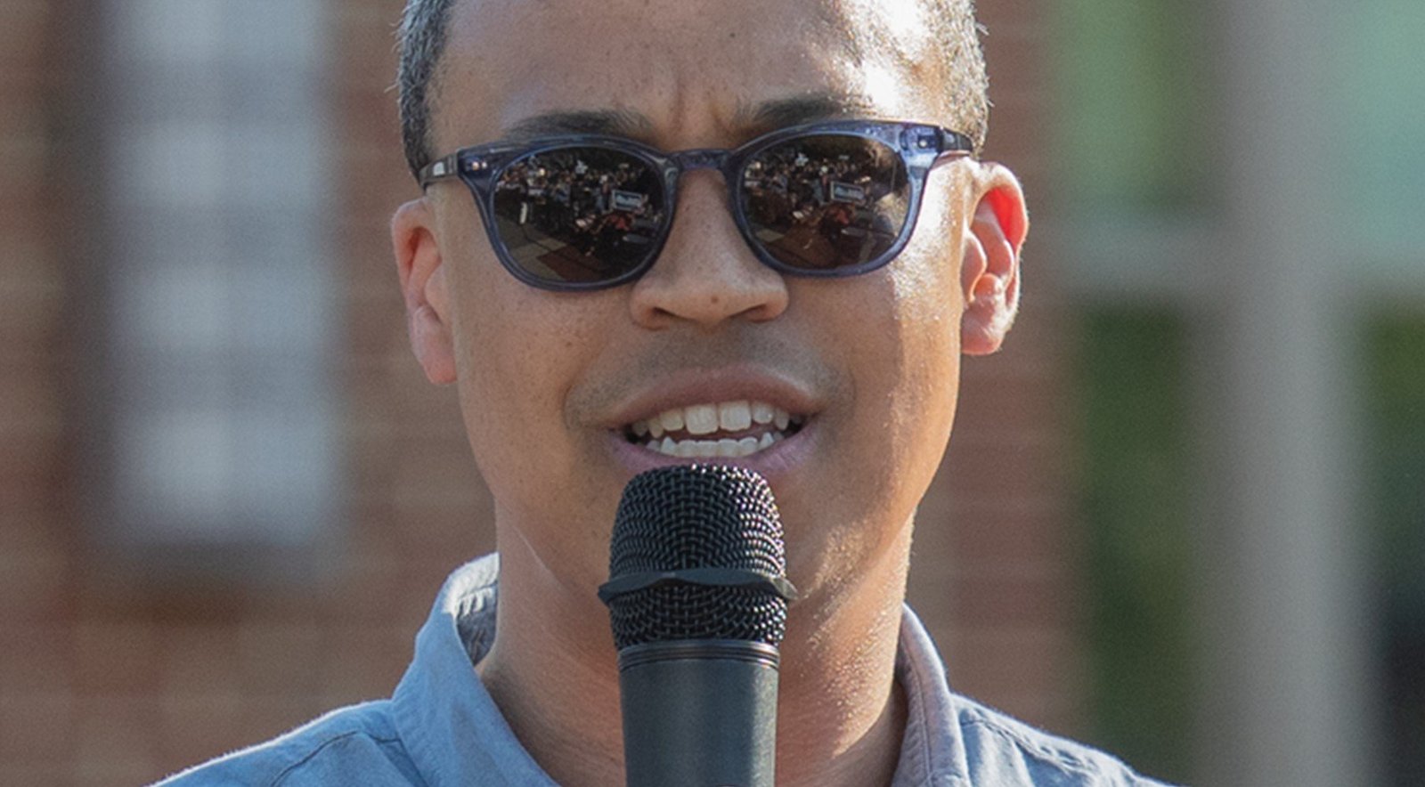 Man speaking into a microphone while wearing sunglasses, with a blurred crowd in the background during an outdoor event.