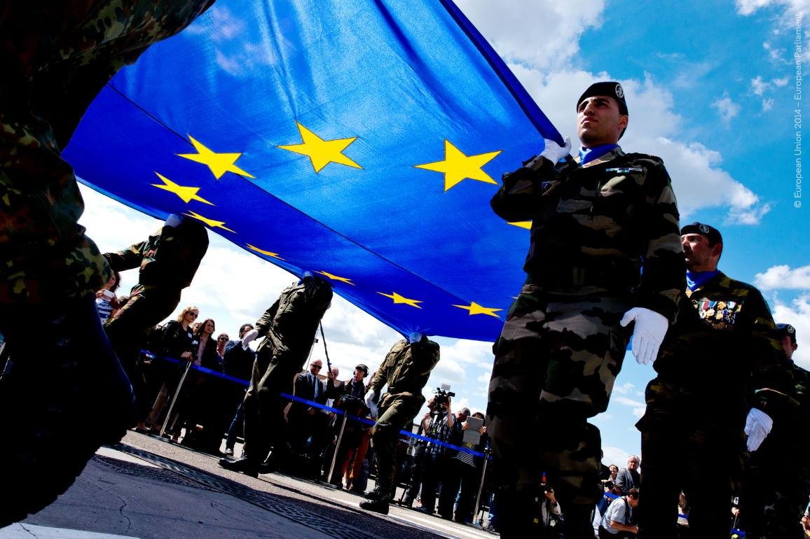 Soldiers carrying a large European Union flag during a public event with an audience in the background under a blue sky.