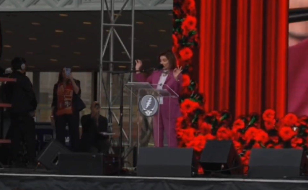 A speaker gestures passionately at a podium adorned with red flowers during an outdoor event, with an audience capturing the moment on their phones.