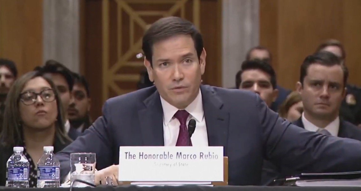 Marco Rubio speaking during a Senate hearing, with attendees listening attentively in the background.