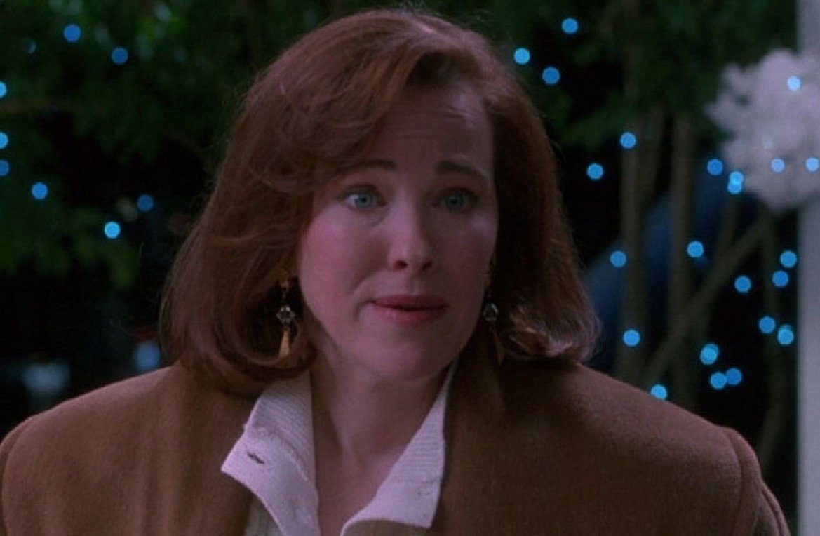 Woman with medium-length brown hair and earrings looks concerned against a backdrop of blue holiday lights, wearing a brown coat over a white sweater.
