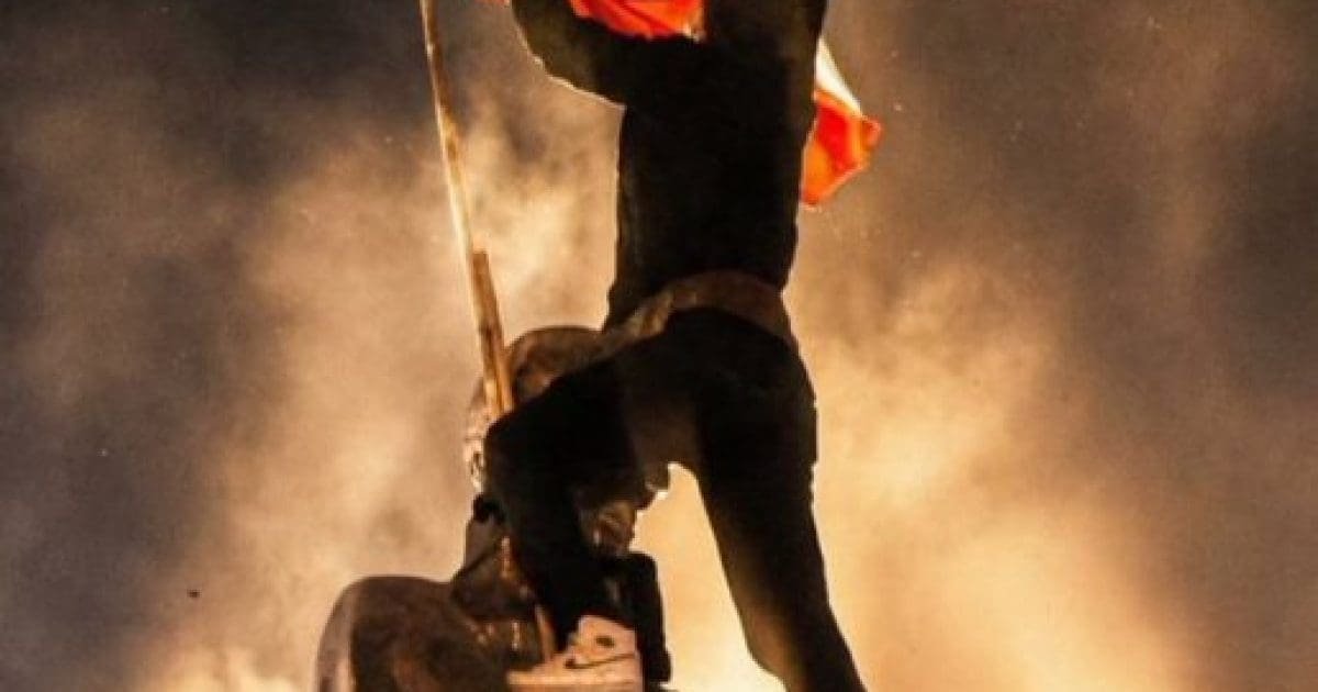 Protester climbing a statue while waving a flag against a backdrop of smoke and light, symbolizing activism and social change.