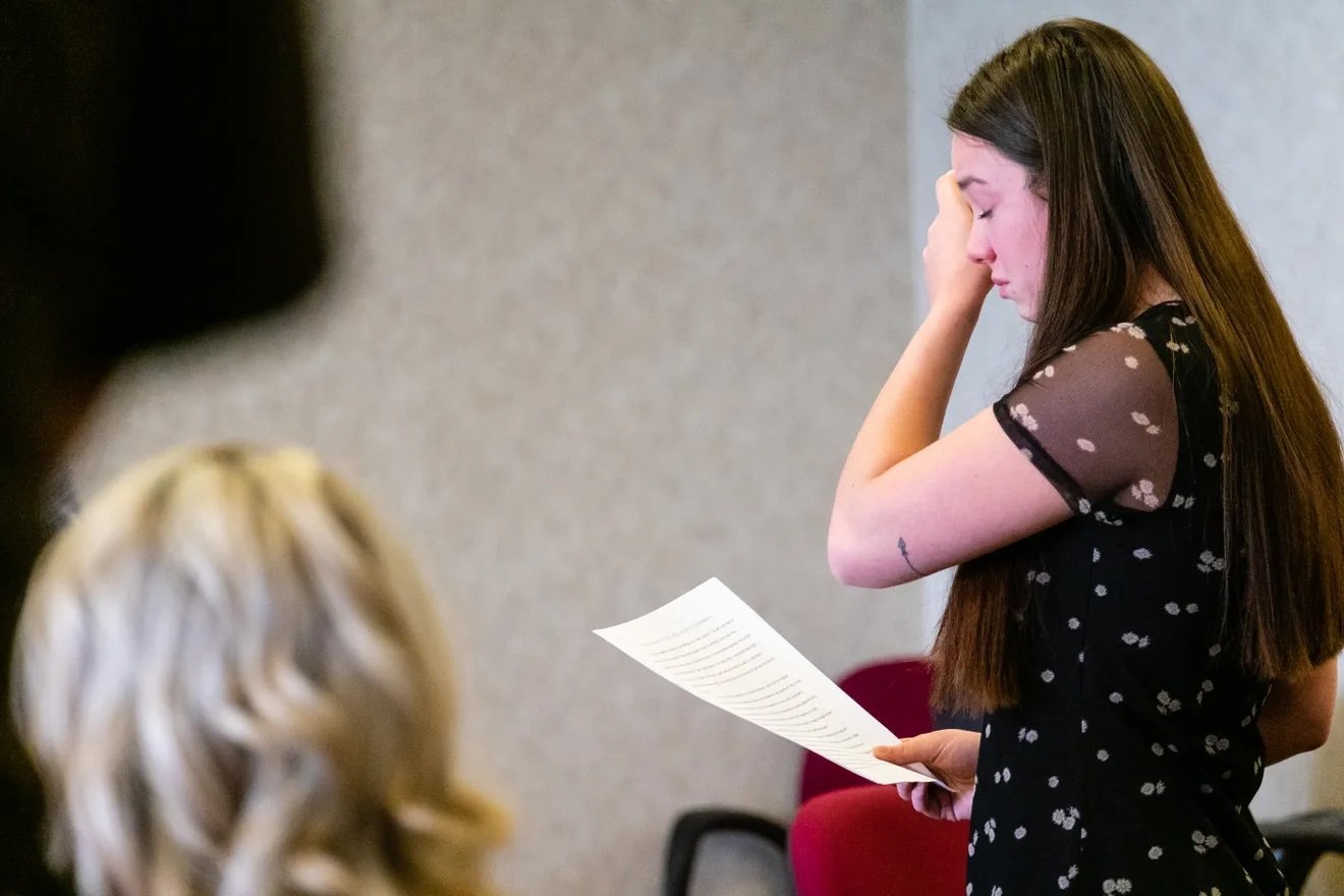 Young woman reading a document while wiping away tears in a courtroom setting, showing emotional distress during a significant moment.
