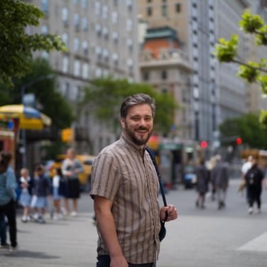 Photograph of a man in a striped, short-sleeved shirt, smiling while standing on a city street
