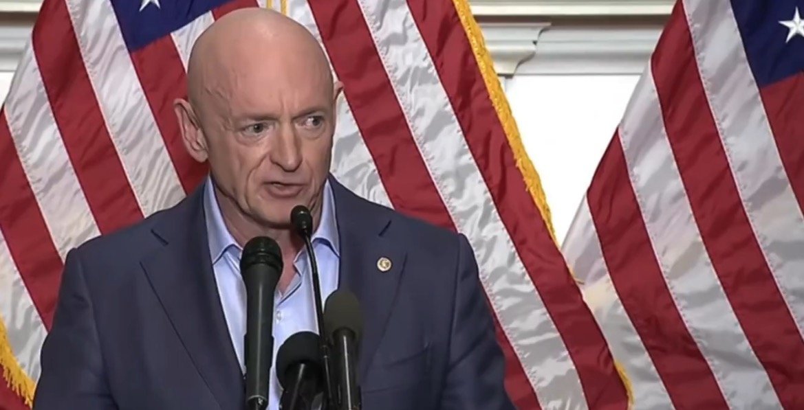 Man speaking at a podium with microphones, in front of American flags, delivering a speech on a public issue.