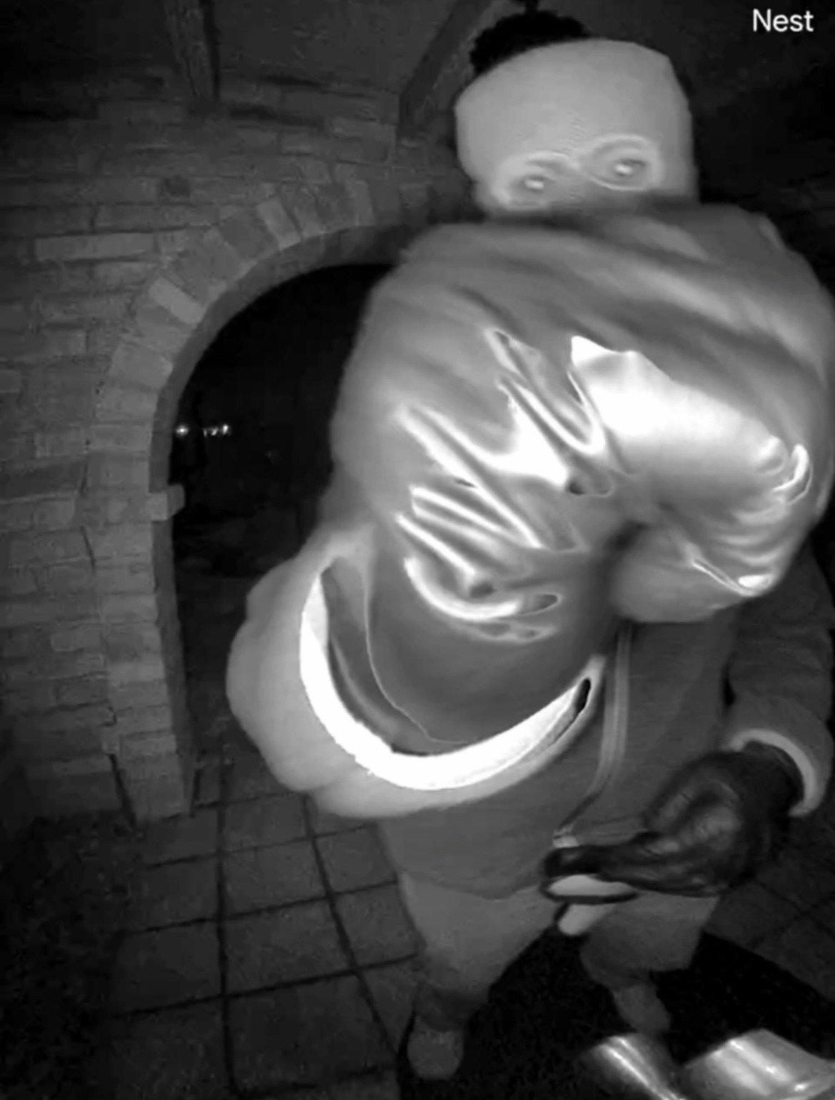 Intruder wearing a beanie and gloves approaches a home at night, holding a boxing glove in a dimly lit entryway.