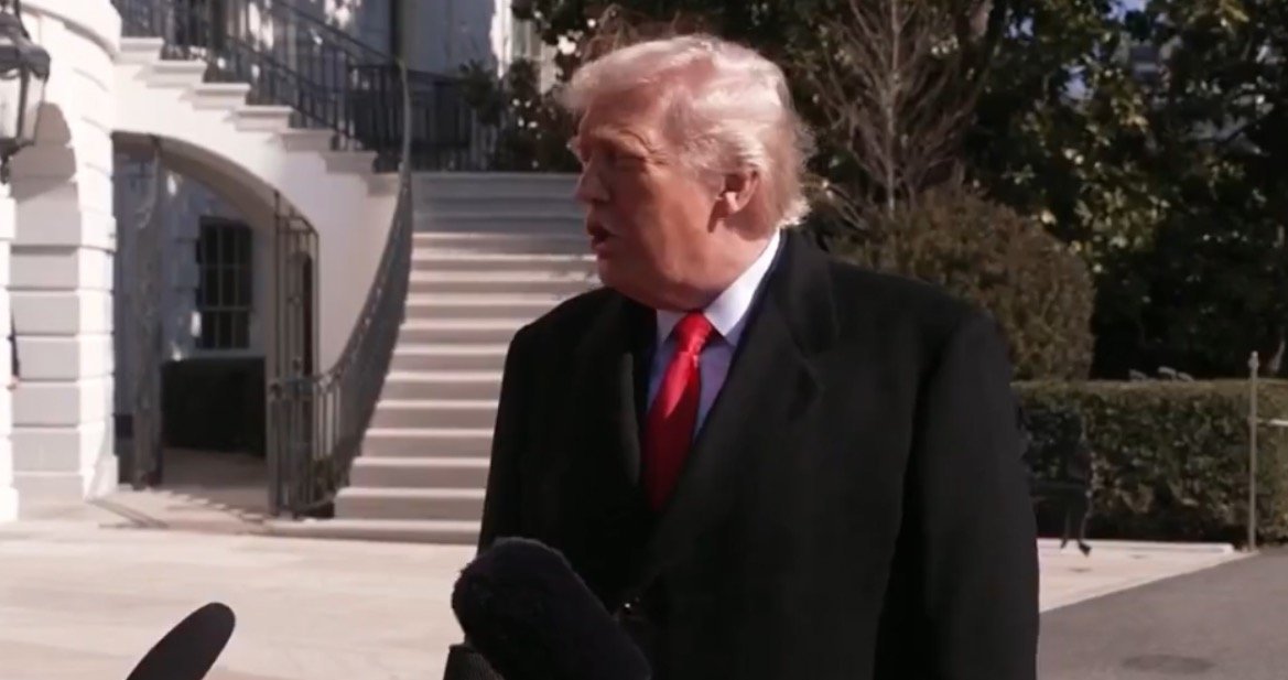 Donald Trump speaking to reporters outside the White House, wearing a black coat and red tie, with a staircase and greenery in the background.