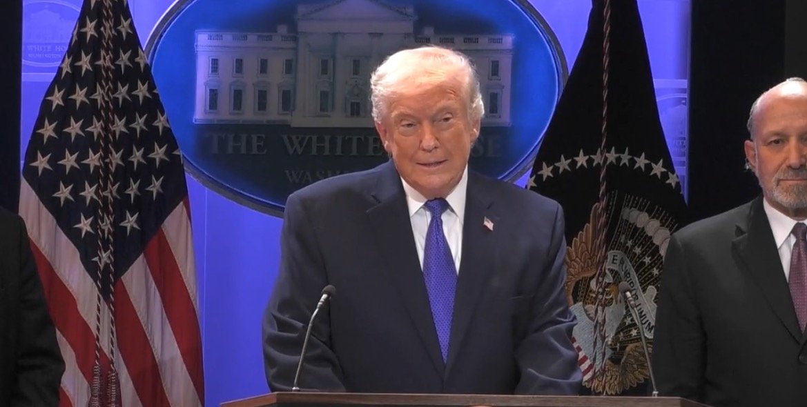 Former President Donald Trump speaking at a press conference in front of the White House, flanked by American flags and an official seal.