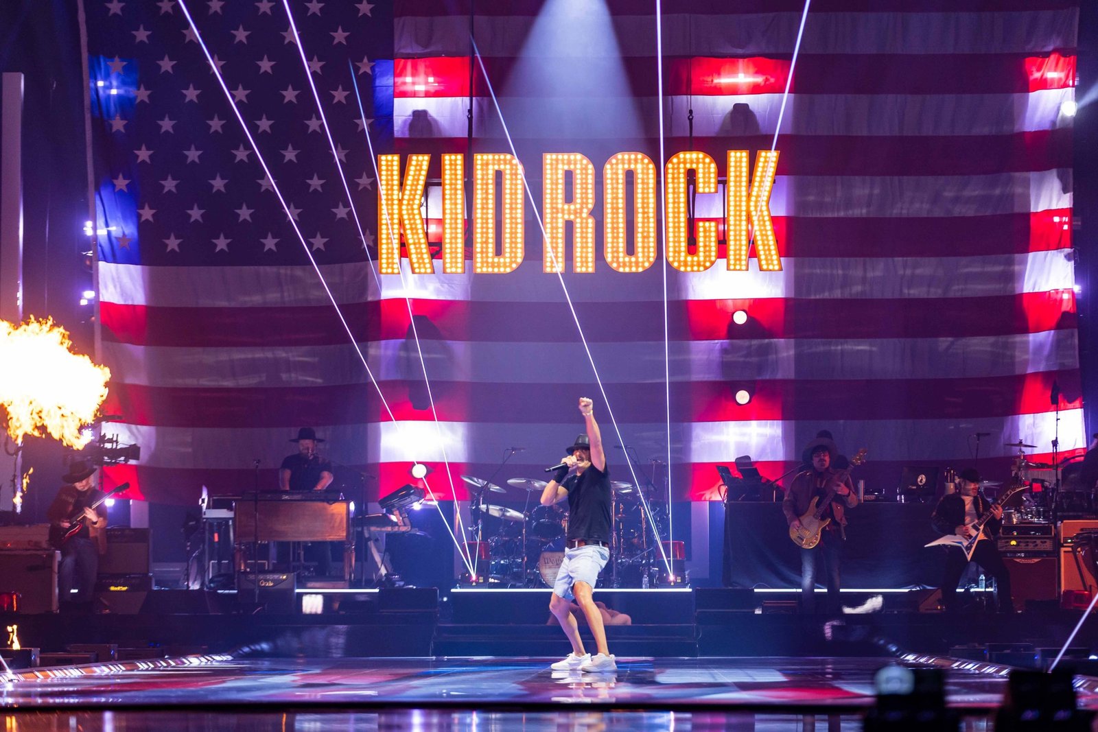 Kid Rock performs on stage with an American flag backdrop, showcasing vibrant lights and energetic musicians during a live concert.