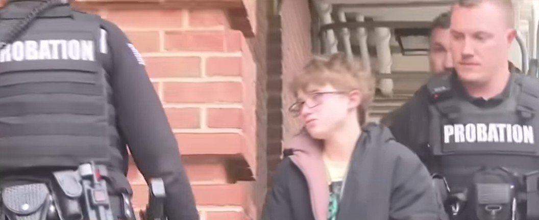 Two probation officers escort a young person wearing glasses outside a building, highlighting a serious situation involving law enforcement.