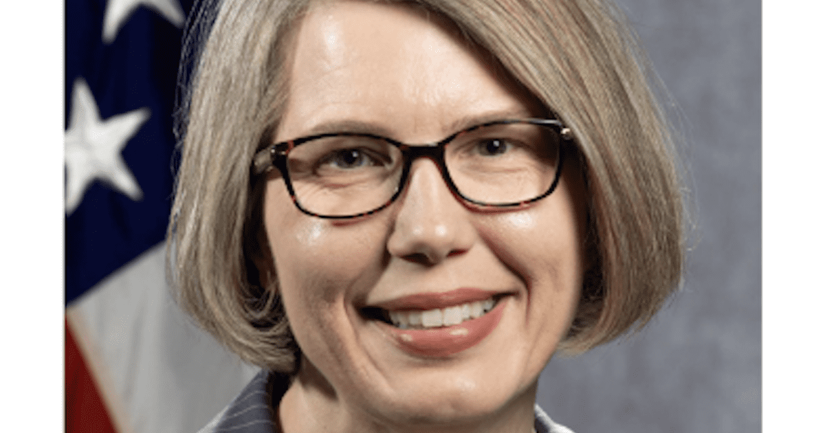 Professional headshot of a woman with short gray hair and glasses, smiling against a blurred background featuring the American flag.