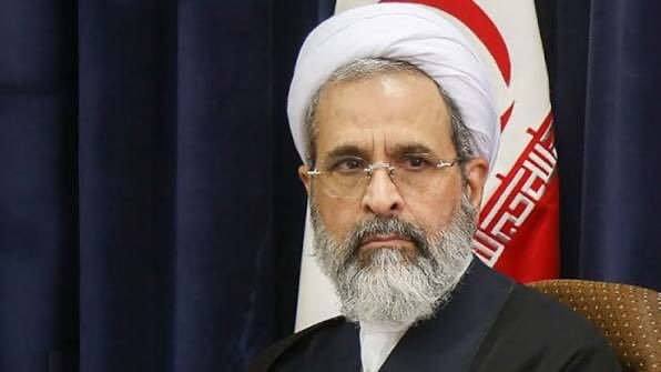 Portrait of a religious leader in traditional attire, seated against a backdrop featuring the Iranian flag.