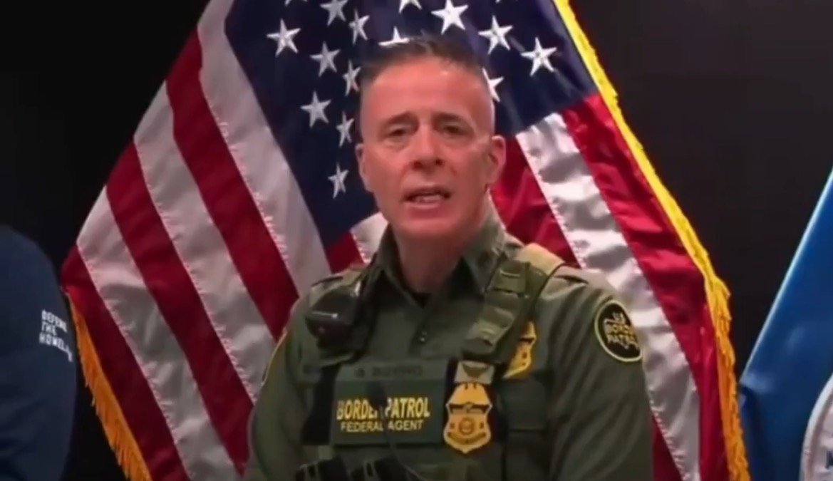 Border Patrol agent speaking at a press conference with the American flag in the background, highlighting law enforcement efforts.