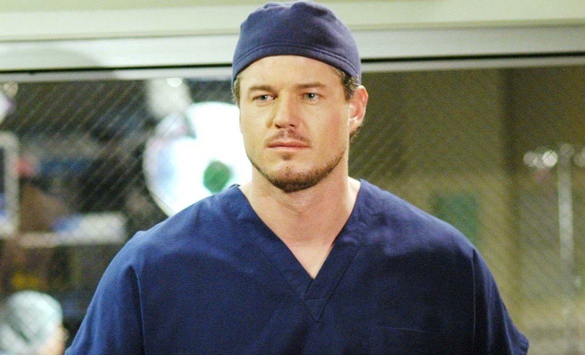 Male surgeon in blue scrubs and cap, looking serious in a hospital setting, with medical equipment blurred in the background.
