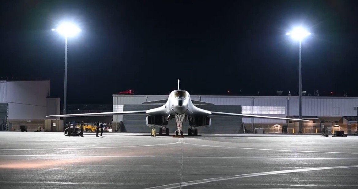 A military aircraft on a tarmac at night, with personnel and equipment visible under bright lights.