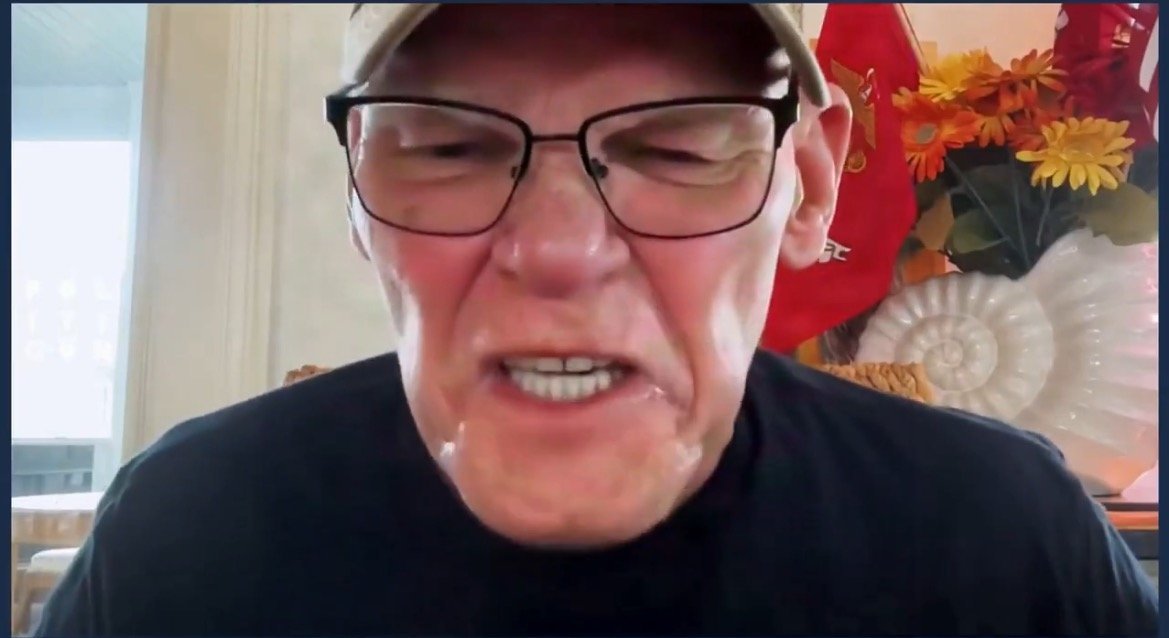 Angry man with glasses expressing strong emotions during a video call, with decorative flowers and flags in the background.