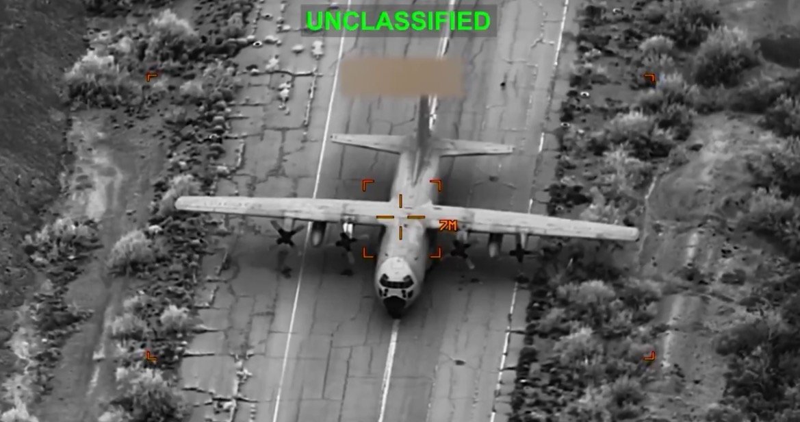 Military aircraft captured from an aerial perspective, displaying its features on a cracked road surrounded by vegetation, marked as unclassified.