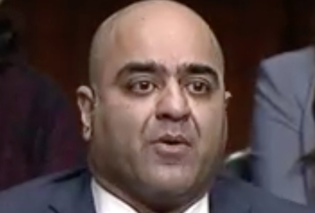 Man with a bald head and suit speaking during a formal event, expressing concern or making a statement, with a focused expression and audience in the background.