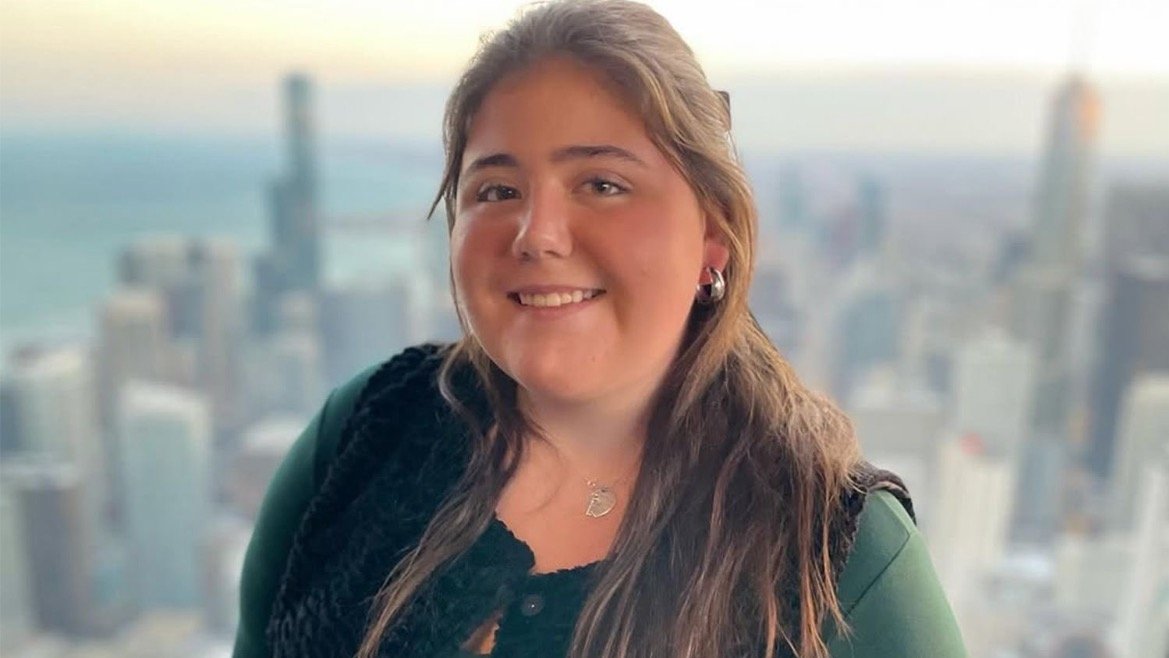 Smiling young woman with long hair, wearing a green top and black vest, poses against a city skyline background.