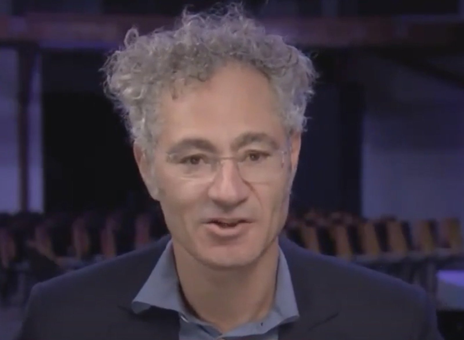 Man with curly hair and glasses speaking in a professional setting, conveying insights during a presentation or interview.
