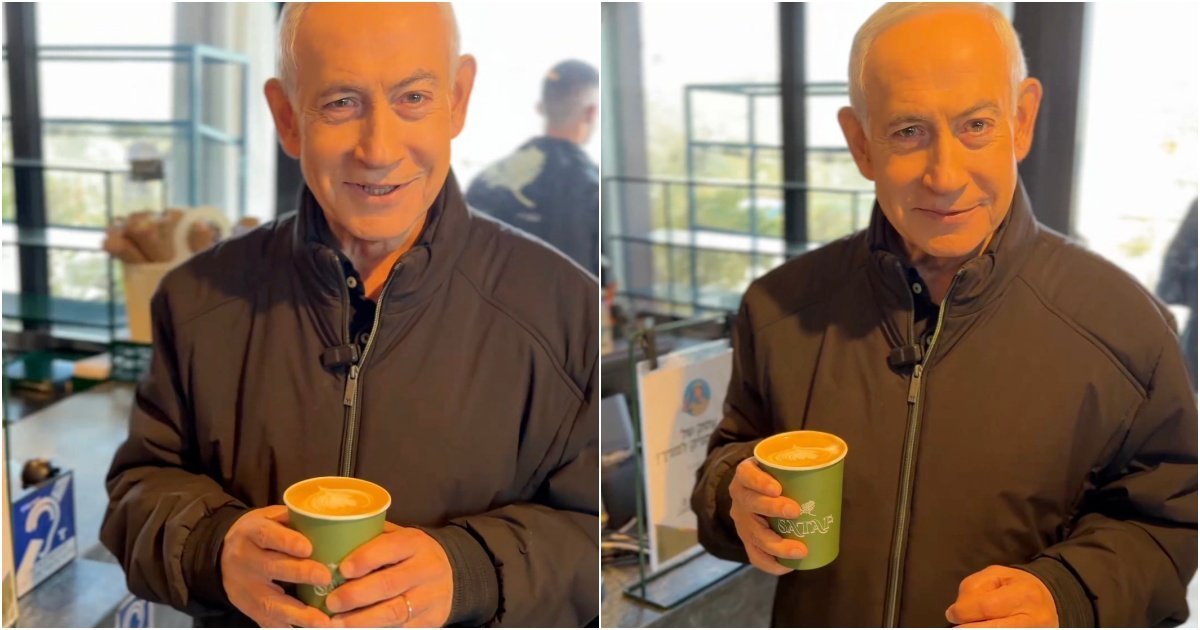 Man in a black jacket holding a coffee cup, smiling while standing in a café with a modern interior and glass display.