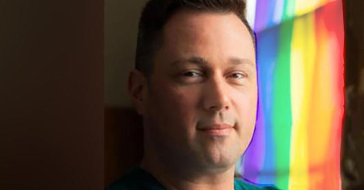 Portrait of a man with a neutral expression, standing near a colorful rainbow backdrop, highlighting diversity and inclusion themes.