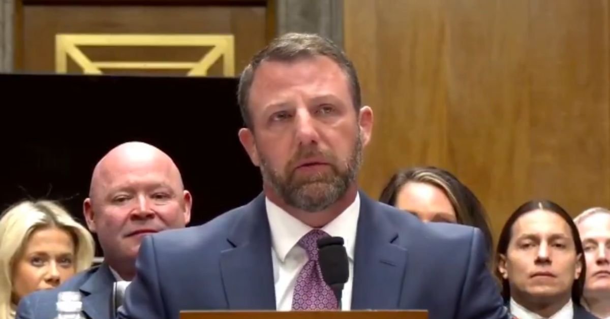A man in a suit speaks at a congressional hearing, with attentive audience members visible in the background.