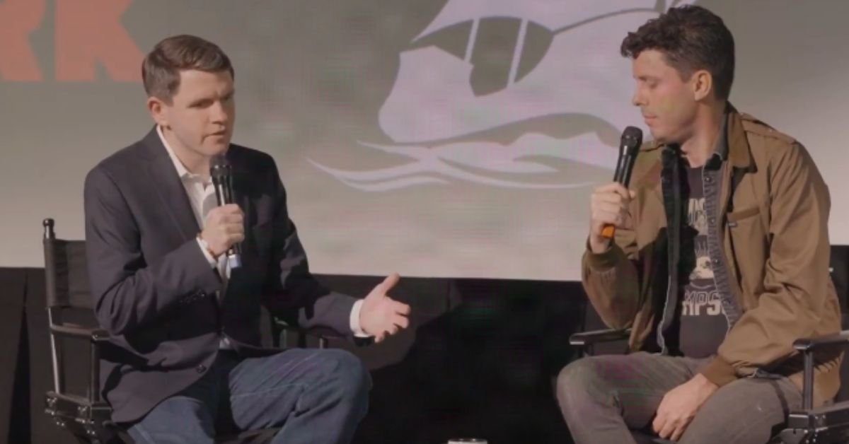 Two speakers engage in a discussion on stage, holding microphones, with a backdrop featuring a ship logo.