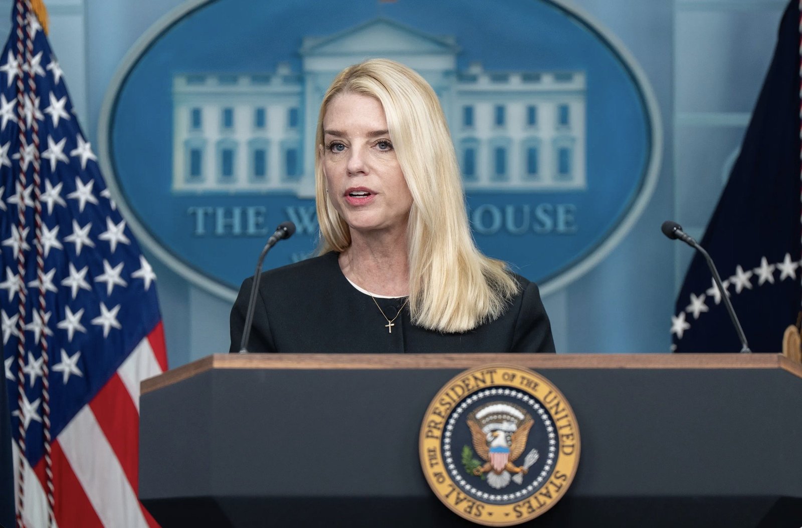 Press briefing at the White House featuring a female speaker in a black outfit, with the presidential seal and American flags in the background.