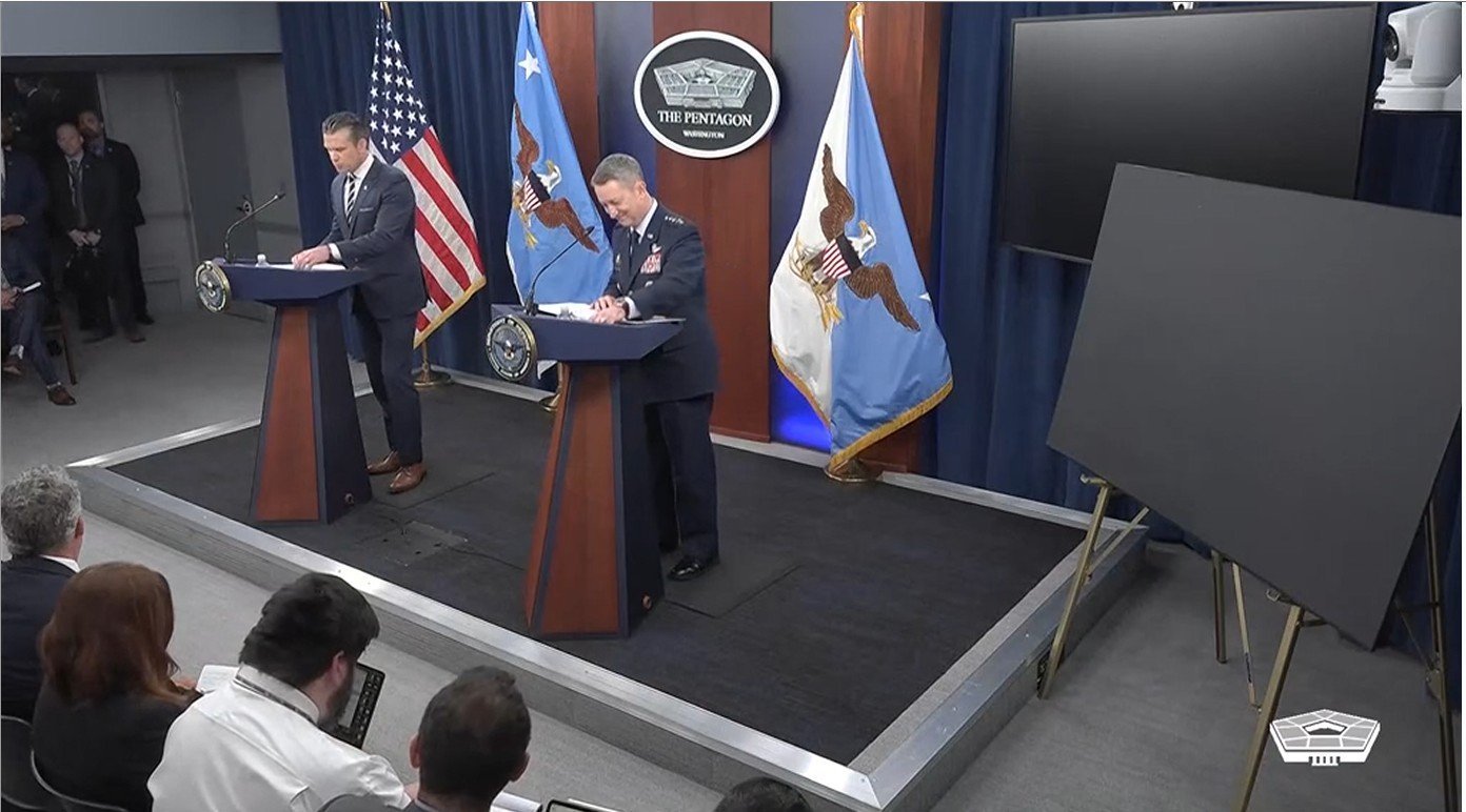Pentagon press briefing featuring military officials at podiums, with American flags and media members in attendance.