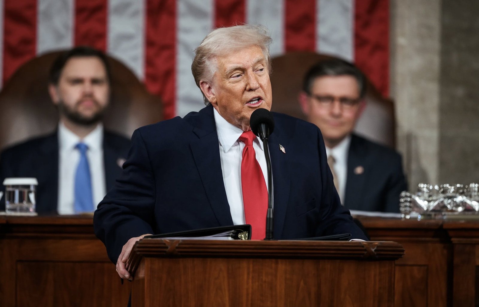 Donald Trump delivers a speech at the podium in front of an American flag backdrop during a congressional address, with two officials seated behind him.