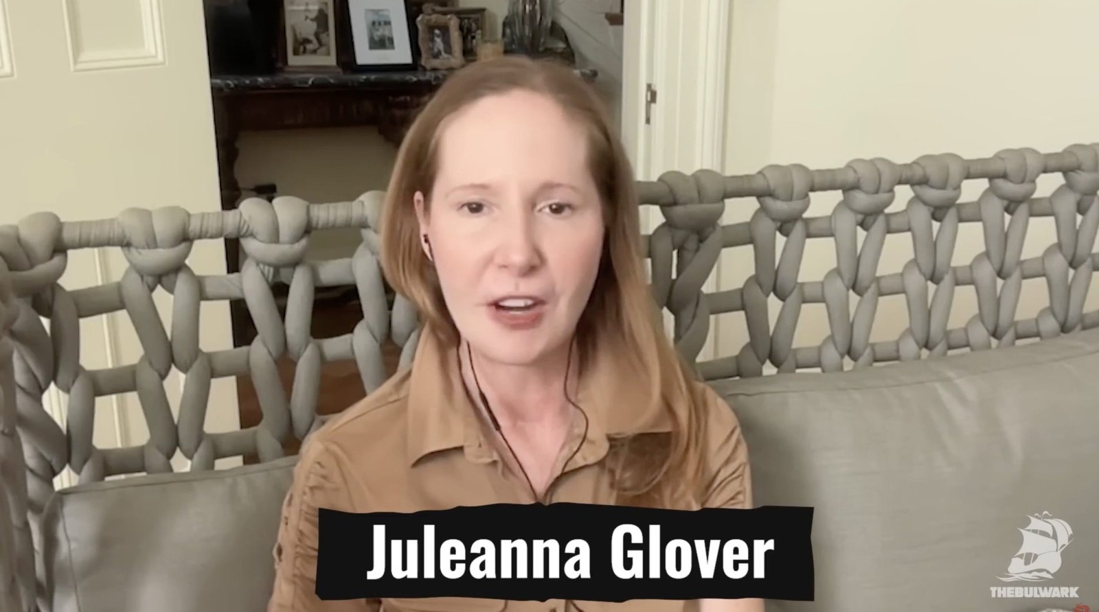 Juleanna Glover speaks during a virtual interview, showcasing a modern interior with a unique woven chair design in the background.