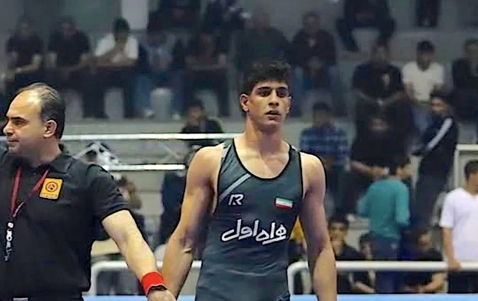 Young male wrestler wearing a green uniform stands with a referee in a competitive wrestling event, surrounded by an audience in the background.