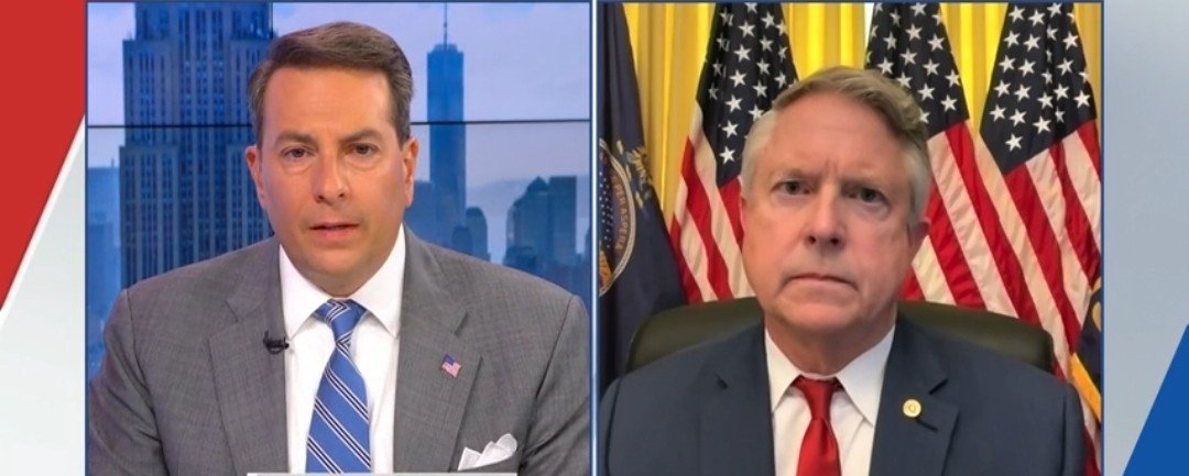 Two news anchors discussing current events, with a backdrop of the New York City skyline and American flags, during a televised interview.