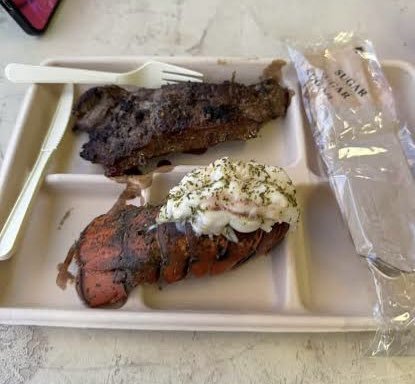 Grilled steak and lobster tail with garlic butter served on a tray, accompanied by a sugar packet and plastic utensils.