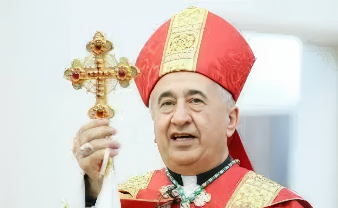 A religious leader in a red ceremonial robe holds a ornate cross, delivering a message during a formal event.